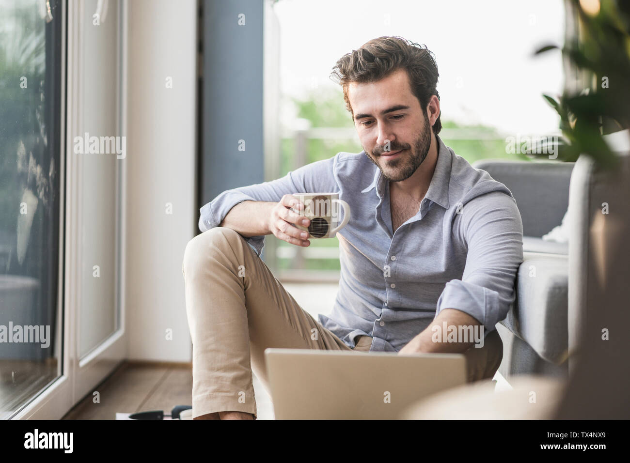 Giovane uomo seduto a casa sul pavimento, utilizzando laptop, bere caffè Foto Stock