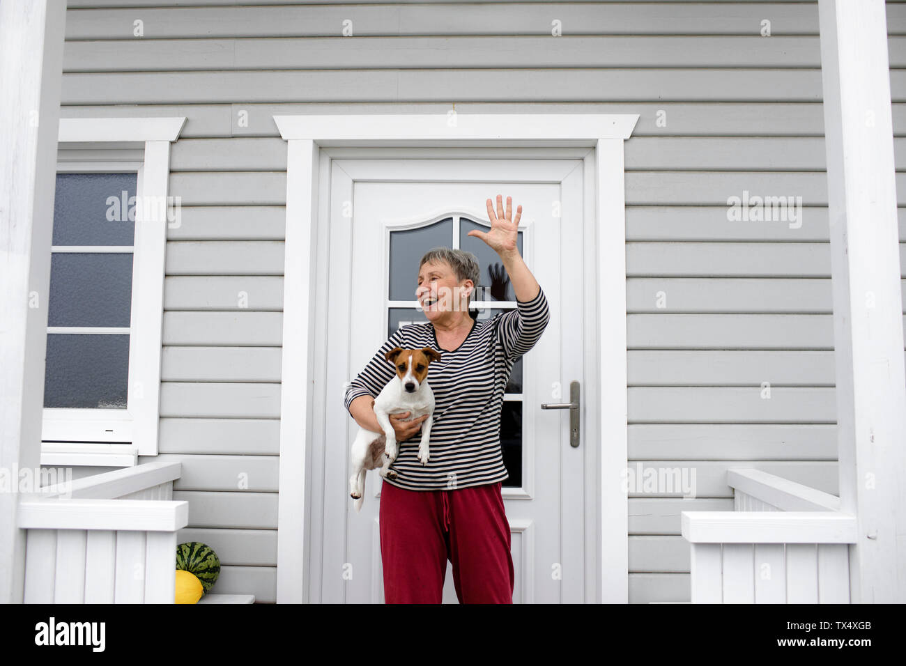 Felice donna senior sul portico con il suo cane alzando la mano Foto Stock