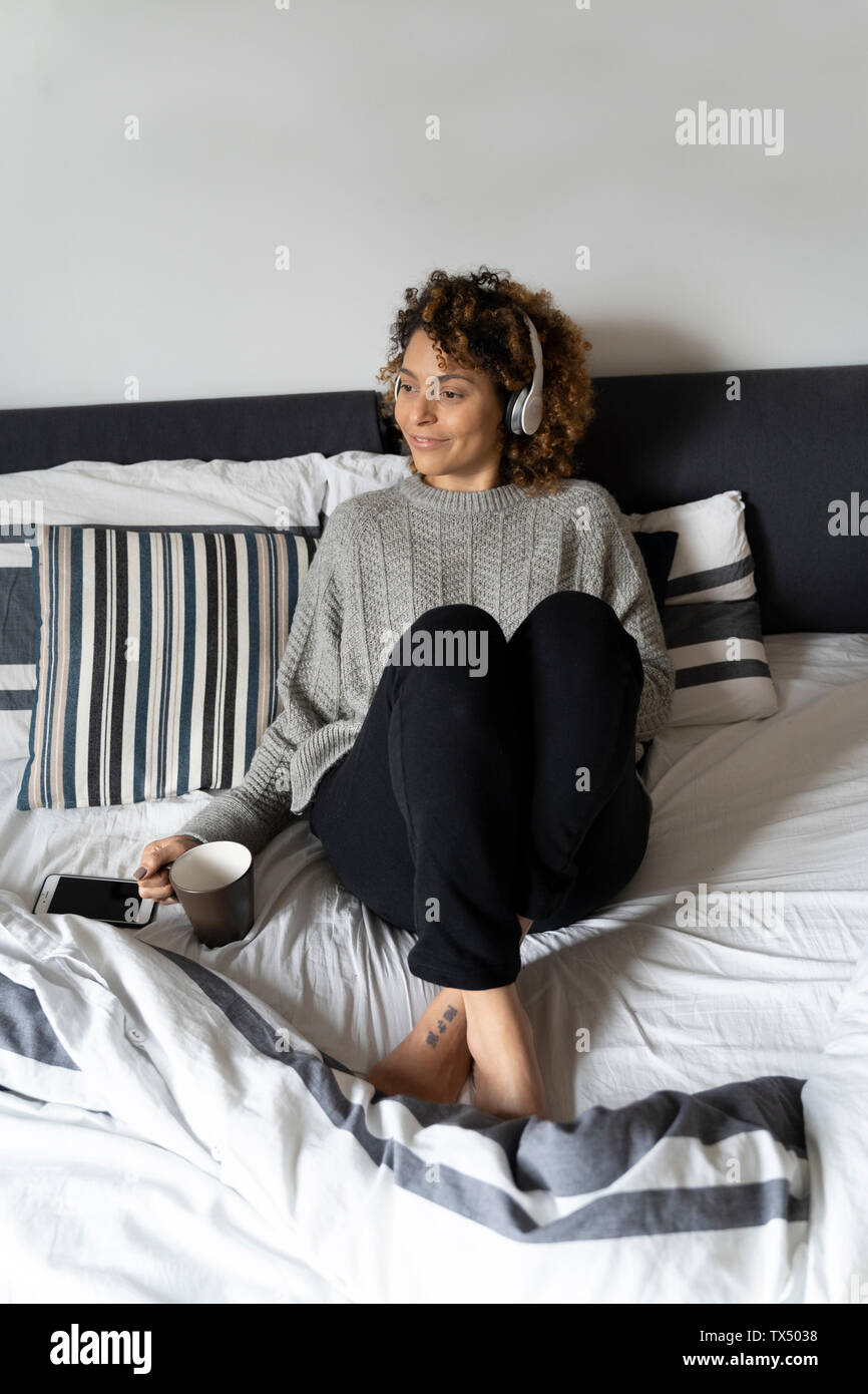 Donna seduta sul letto, bere caffè, ascoltando musica con le cuffie e lo smartphone Foto Stock