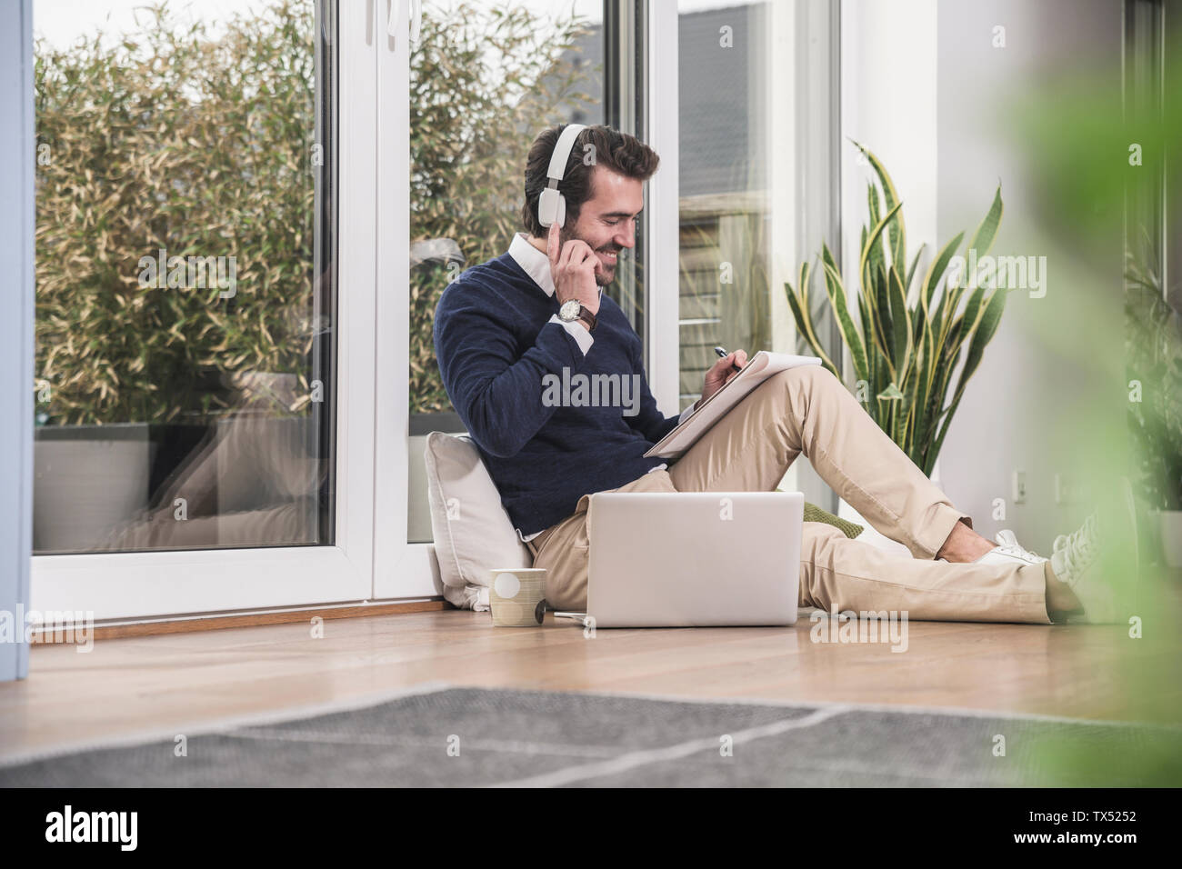 Giovane uomo seduto in soggiorno, poggiando sulla finestra, lavorando con il computer portatile, prendendo appunti Foto Stock
