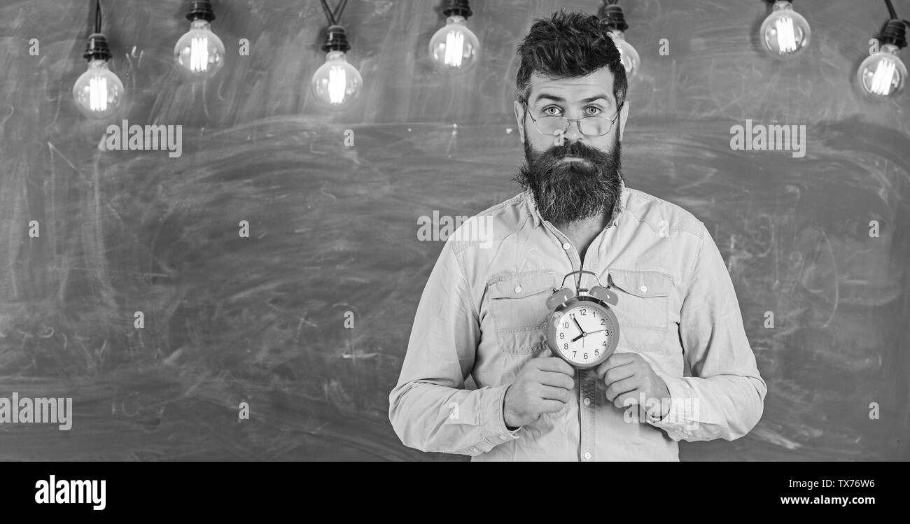 Orario Scolastico concetto. Uomo con barba e baffi sulla faccia calma stand in aula. Barbuto hipster detiene orologio, lavagna su sfondo, copia dello spazio. Insegnante in occhiali tiene sveglia. Foto Stock