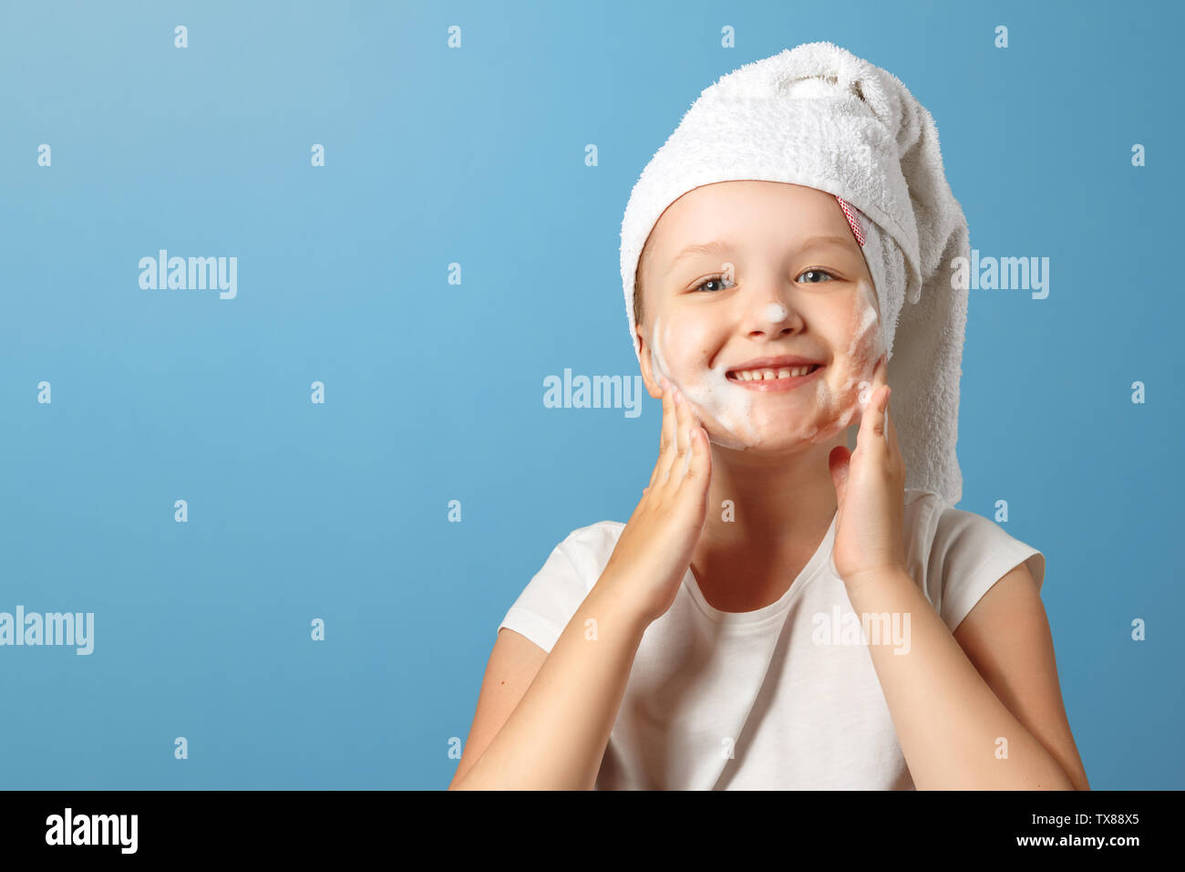 Bambina con un asciugamano bianco sul suo capo lavaggi su uno sfondo blu. Il bambino ride e utilizza la formazione di schiuma sul viso. Il concetto di igiene quotidiana. Foto Stock
