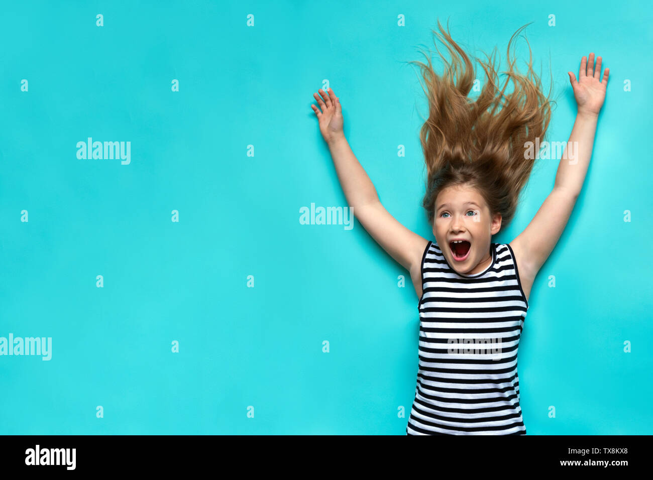 Bambino felice posa su sfondo blu con le mani. Sorridente, ragazza ridere divertirsi vista dall'alto. Infanzia momenti nella vita. Avente un grande tempo. Gioiosa kid in abito a righe vista dall'alto. Carta da parati semplice Foto Stock