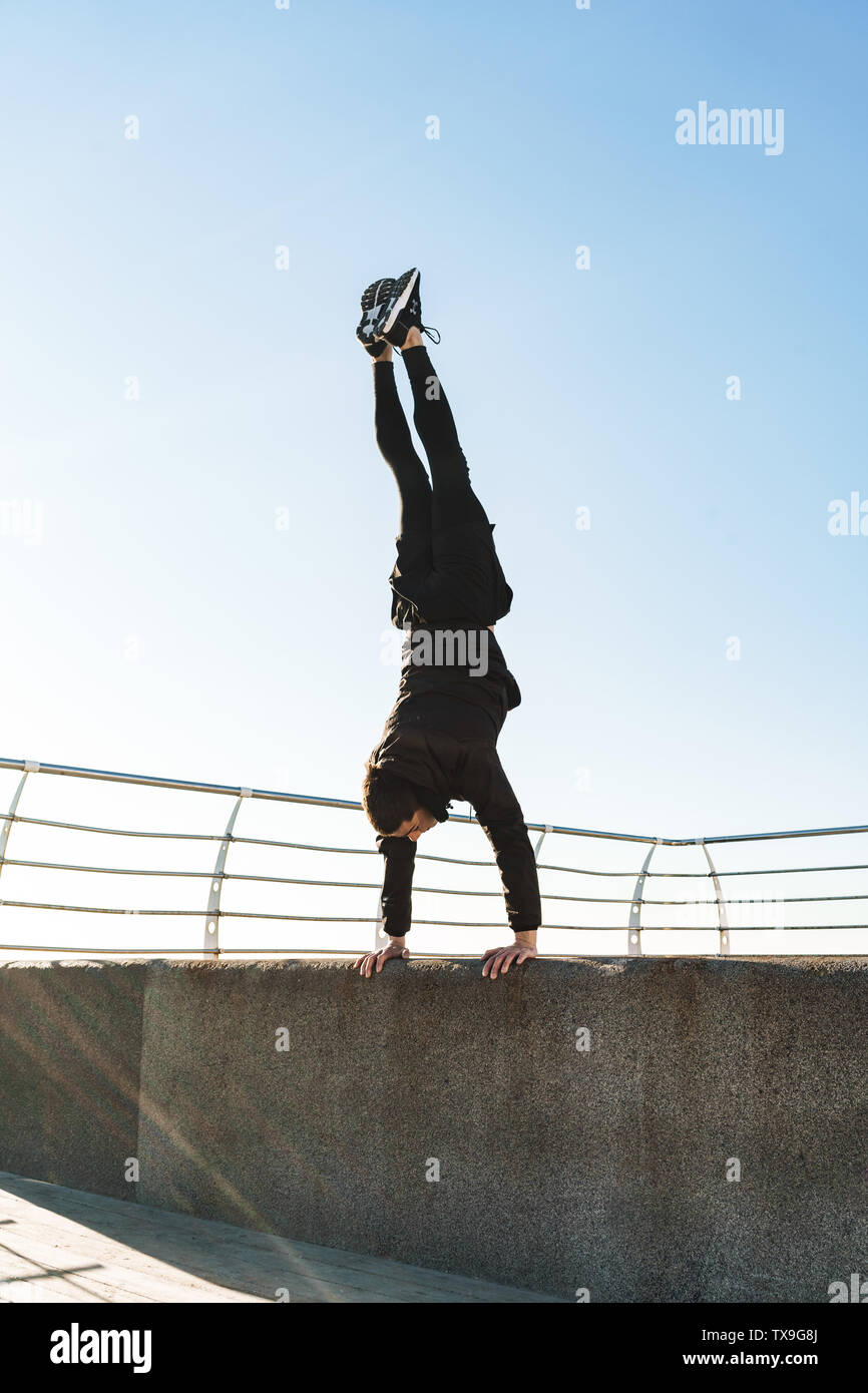 Foto del giovane ragazzo 20s in tuta nera facendo acrobazie e salti durante la mattina allenamento dal mare Foto Stock