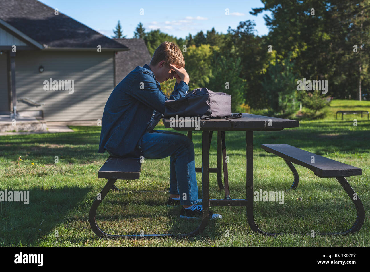 Sconvolto adolescente seduti ad un tavolo da picnic in un parco tutto da solo. Il suo zaino è sdraiato sul tavolo accanto a lui. Foto Stock