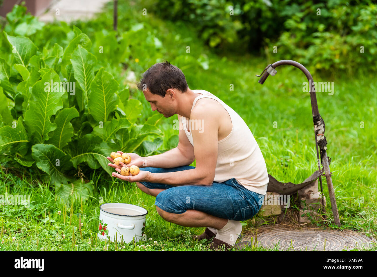 Giovane agricoltore il risciacquo lo sporco da homegrown patate in estate verde in Ucraina seduto da foglie di rafano pianta, ben rubinetto il lavaggio della verdura Foto Stock