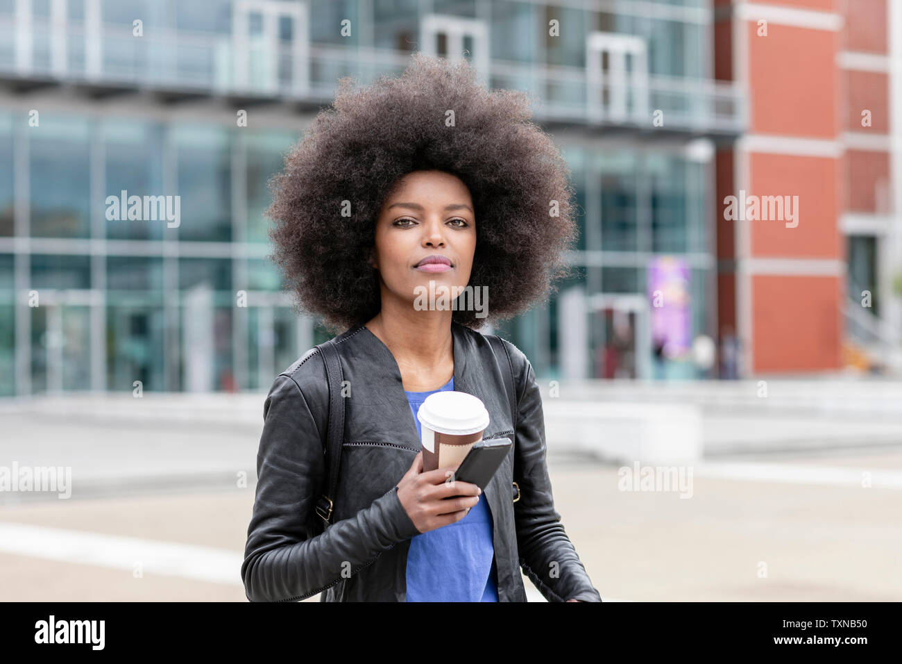 Giovane donna con capelli afro in città, tenendo lo smartphone e il caffè da asporto, ritratto Foto Stock