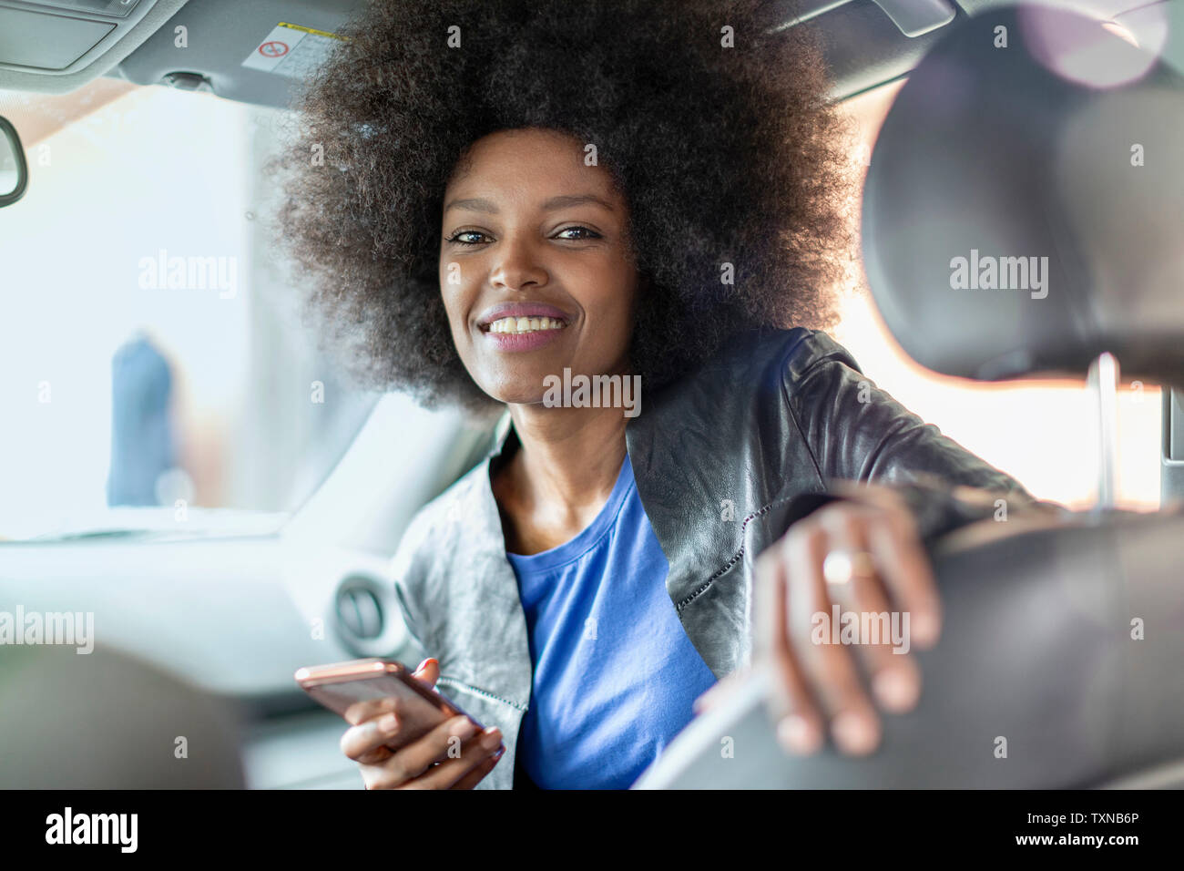 Felice giovane donna con capelli afro in auto sedile passeggero tenendo lo smartphone, ritratto Foto Stock