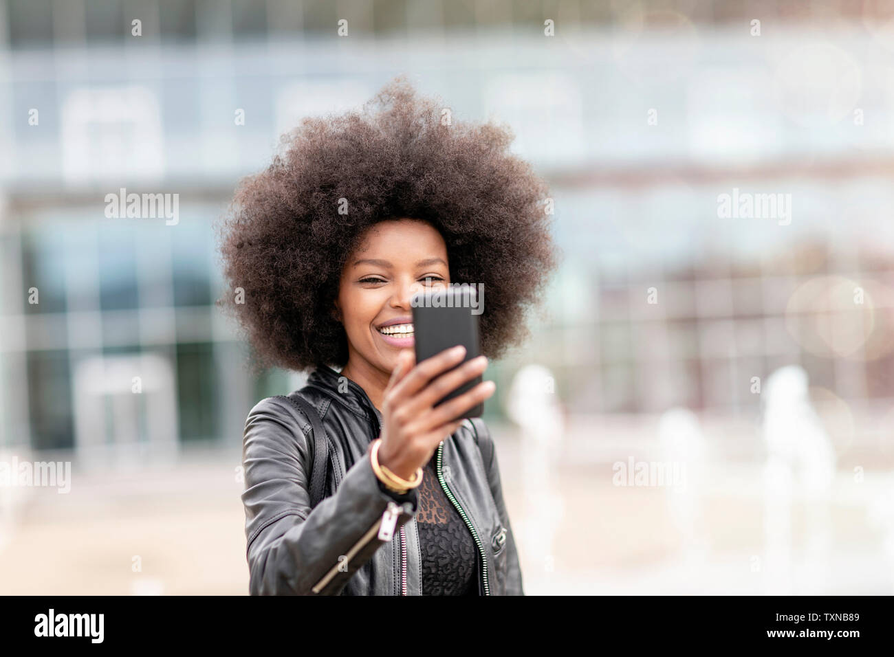 Giovane donna con capelli afro tenendo lo smartphone su selfie atrio della città Foto Stock