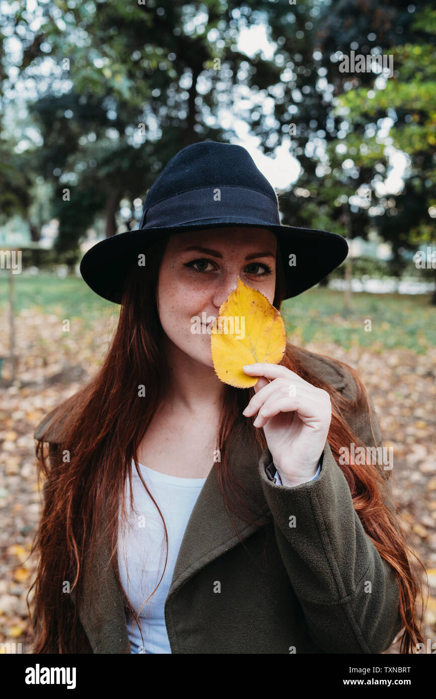 Giovane donna con lunghi capelli rossi holding autumn leaf sul naso in park, ritratto Foto Stock