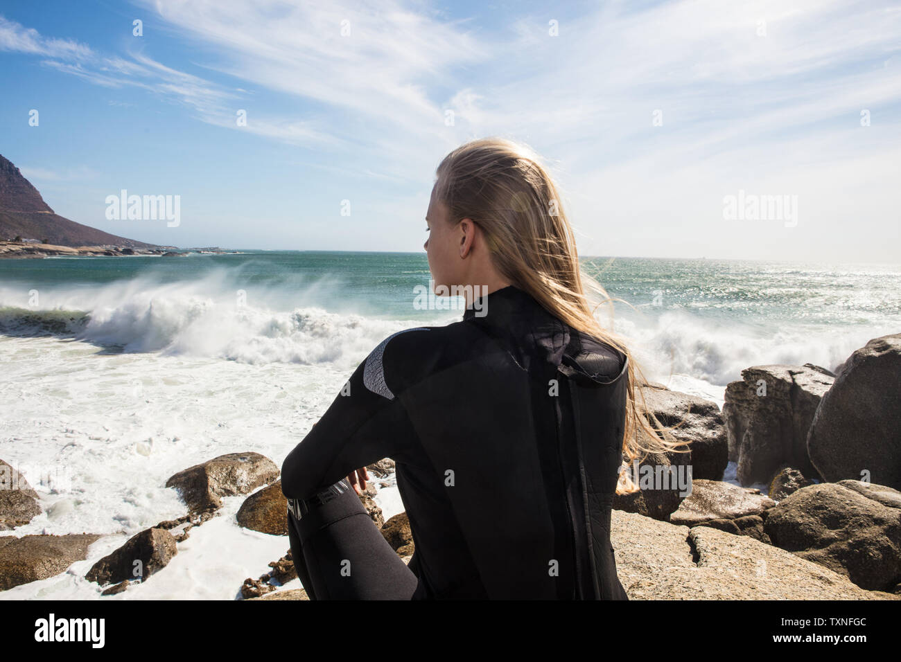 Giovani femmine surfer con lunghi capelli biondi seduto sulla spiaggia rock, vista posteriore, Cape Town, Western Cape, Sud Africa Foto Stock