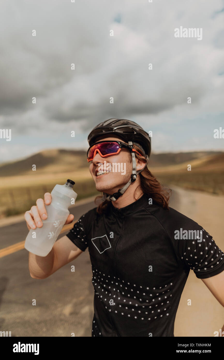 Giovane maschio ciclista tenendo la bottiglia di acqua sulla strada rurale, Exeter, CALIFORNIA, STATI UNITI D'AMERICA Foto Stock