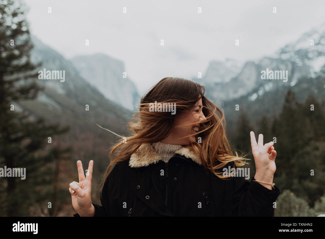 Giovane donna agitando capelli lunghi marrone e facendo segno di pace nel paesaggio di montagna, il Villaggio di Yosemite in California, Stati Uniti d'America Foto Stock