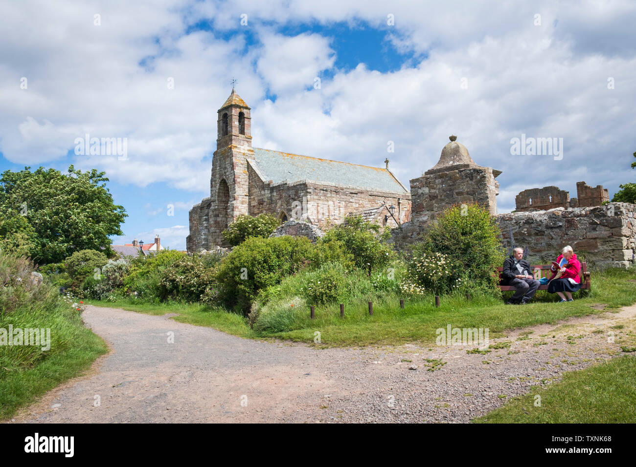 Coppia di anziani sulla giornata fuori in appoggio su una panchina vicino alla chiesa parrocchiale di Santa Maria Vergine su Lindisfarne, Isola Santa, Northumberland Foto Stock
