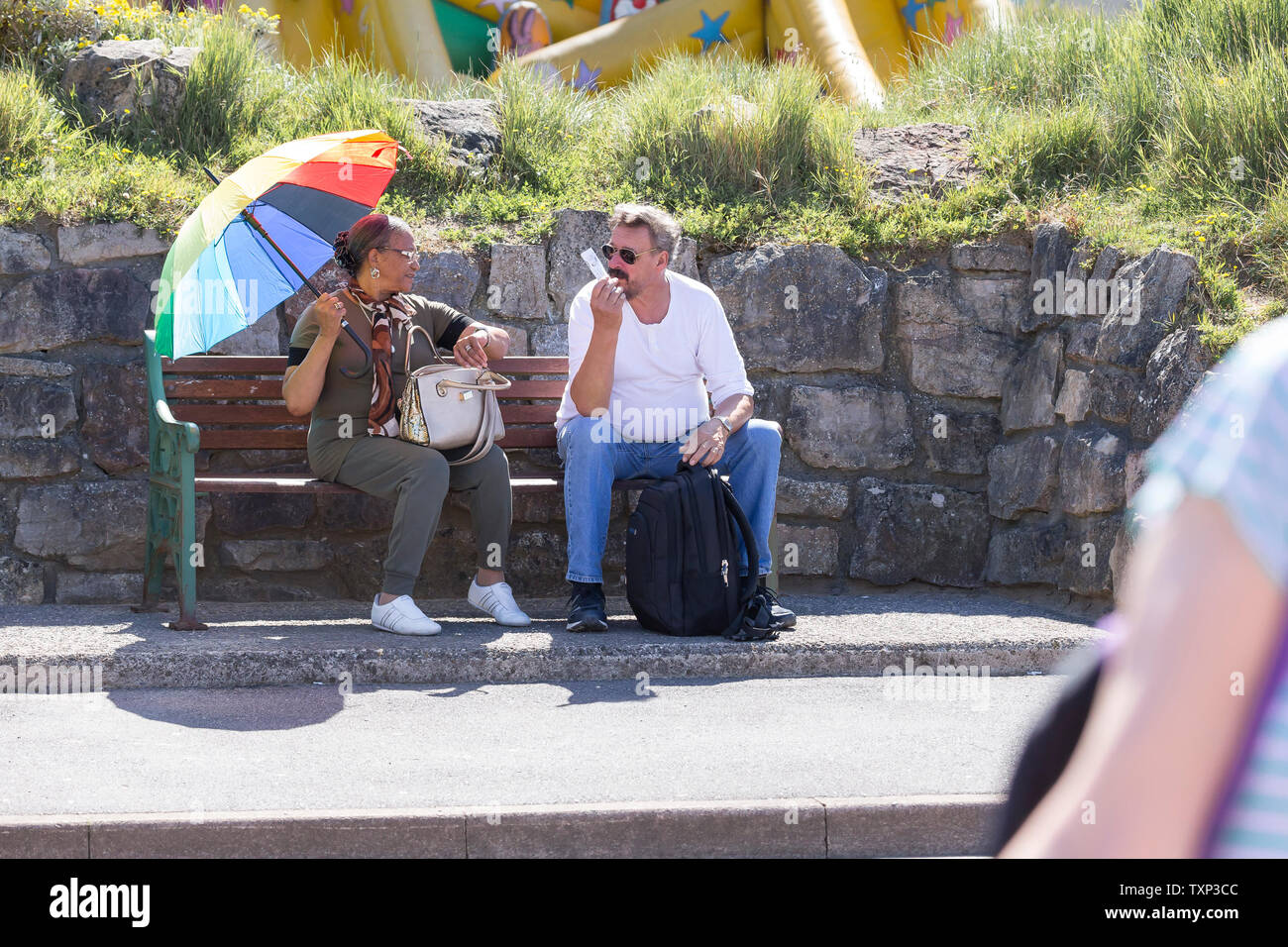 Coppia isolata di mezza età seduta su panca all'aperto a Weston-Super-Mare, Regno Unito, godendo del sole estivo sotto il grande ombrellone multicolore come ombrellone. Foto Stock