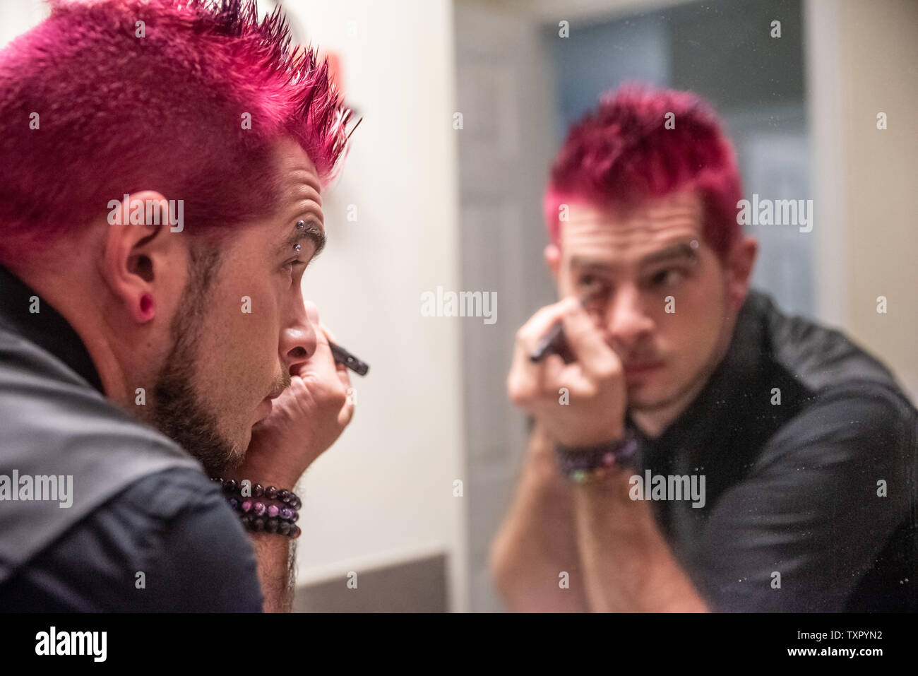Diversi caucasici uomo maschio con dentato per capelli rosa applicazione eyeliner specchio dello specchio di riflessione. Indossa abiti neri con punk rocker l alternativa Foto Stock