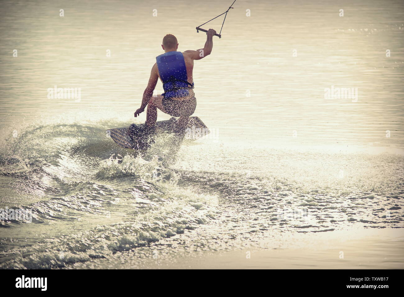 Surfer wakeboarding un Ollie salto al tramonto dorato. Esperto wakeboarder facendo Ollie jump, spruzzi di gocce di acqua nella fotocamera. Giovane uomo che cavalca c Foto Stock