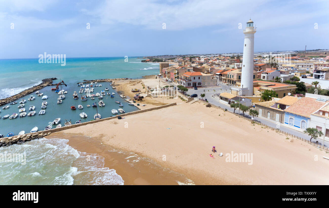 Spiaggia di sabbia dorata con acque poco profonde, piccolo porto con barche, bianco di altezza faro in siciliano di villaggio di pescatori di Punta Secca, set di serie TV Insp Foto Stock