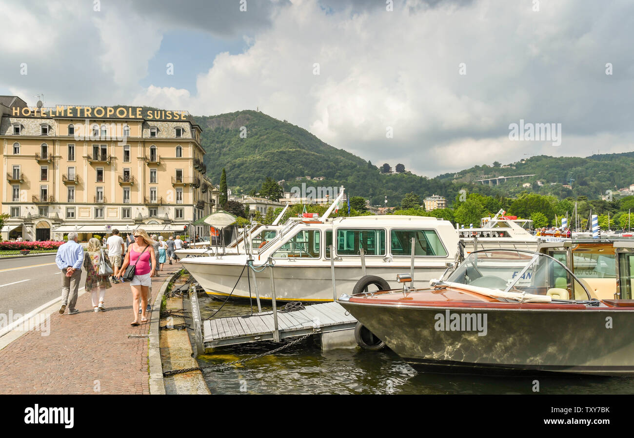 COMO, Italia - Giugno 2019: persone passeggiate lungolago con barche a motore accanto a Como sul Lago di Como. In fondo è l'Hotel Metropole Suisse Foto Stock
