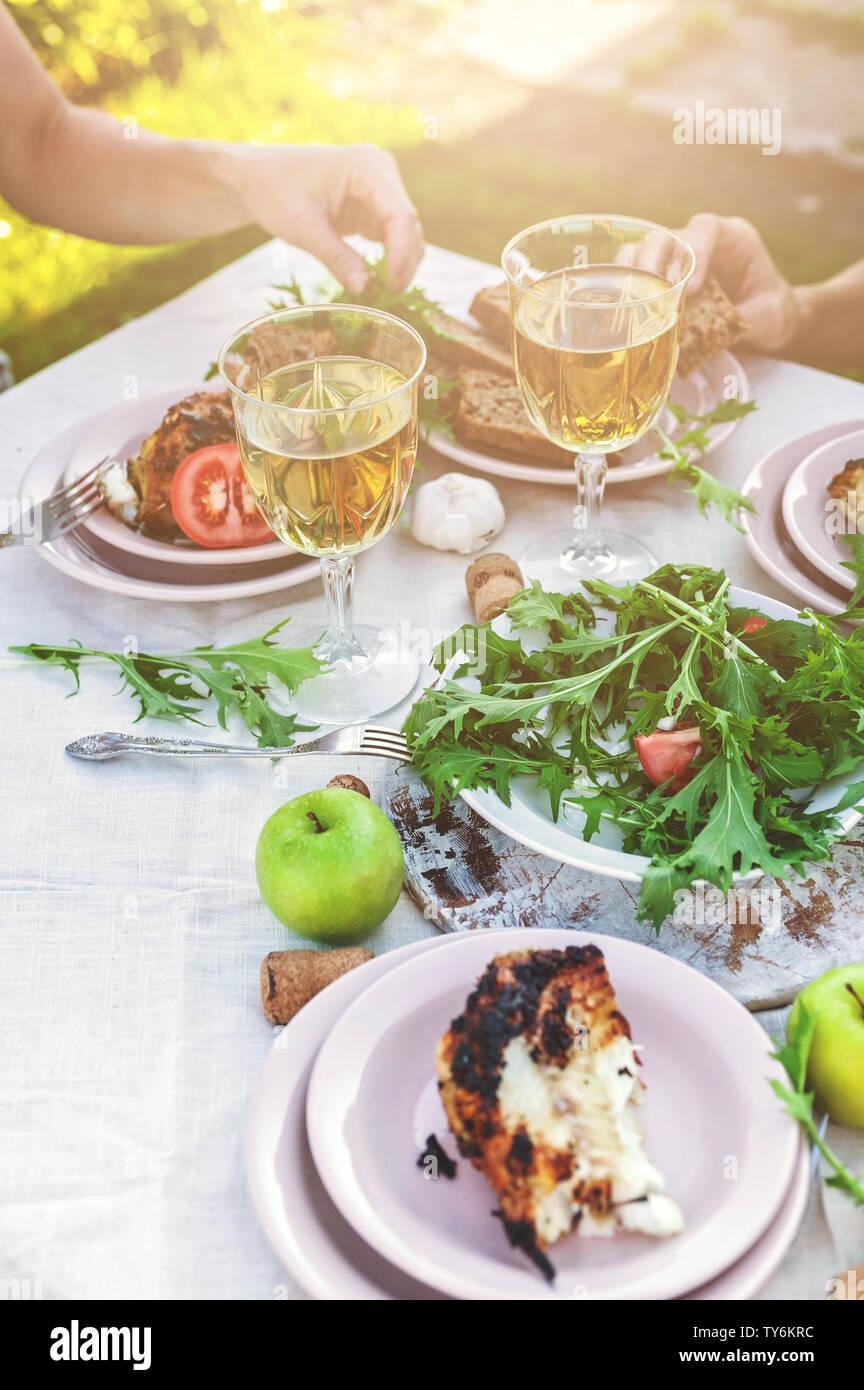 La gente mangia al tavolo in giardino. La cena al tramonto con vino e grigliate di pesce, verdure fresche e erbe aromatiche. Colpo verticale Foto Stock