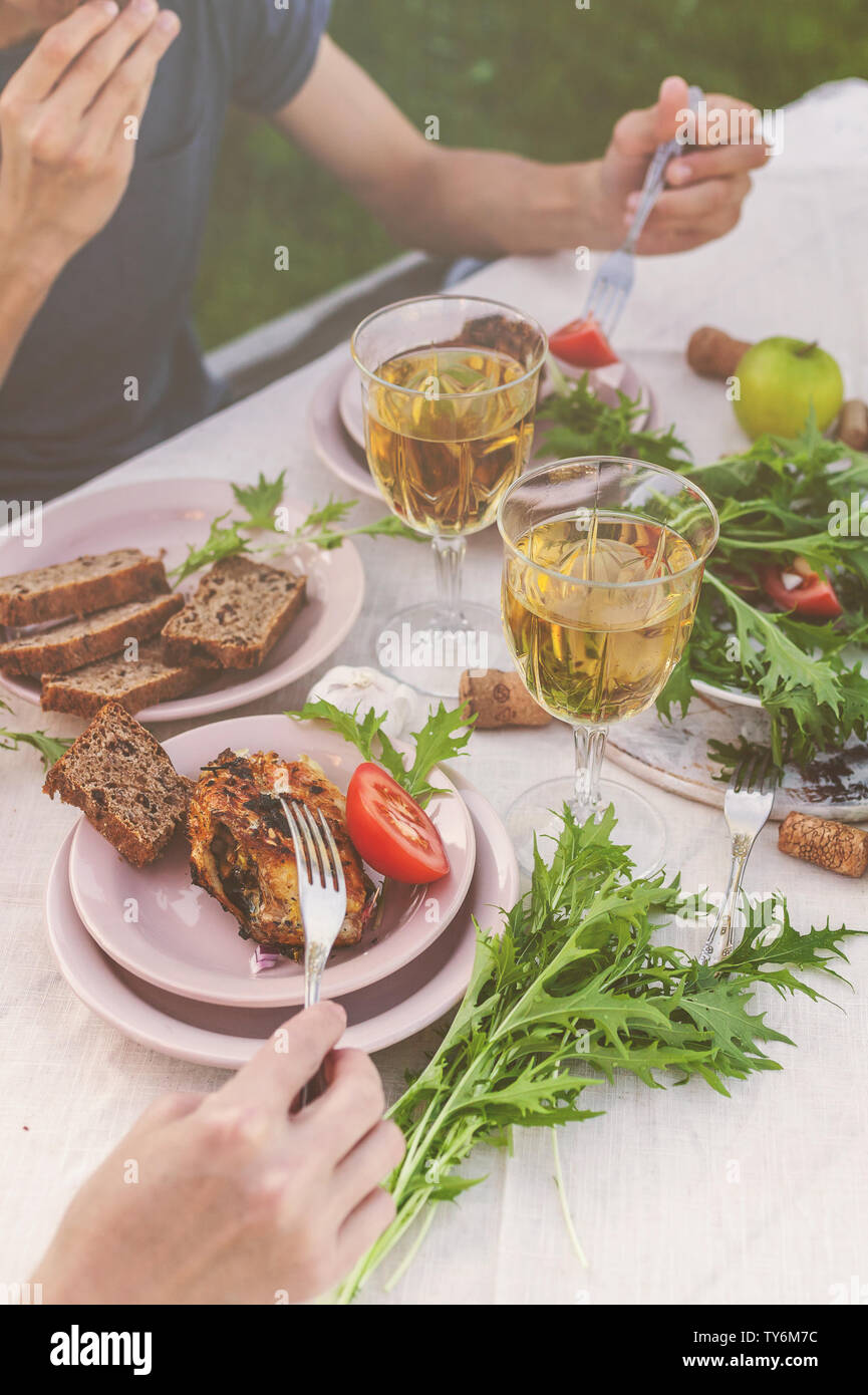 La gente mangia al tavolo in giardino. La cena al tramonto con vino e grigliate di pesce, verdure fresche e erbe aromatiche. Colpo verticale Foto Stock