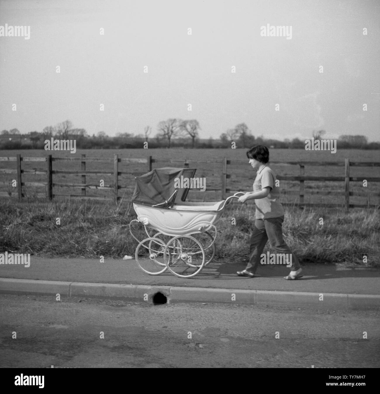 Anni sessanta, storico, una madre spingendo un tradizionale coach-costruito passeggino o carrozzina lungo un marciapiede nel paesaggio rurale, Cossington, Inghilterra. Foto Stock