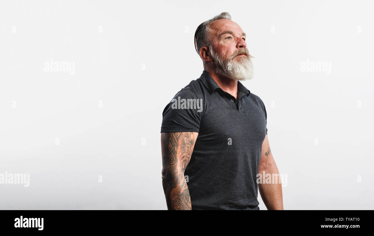 Senior uomo con tatuaggio su bracci in piedi contro uno sfondo bianco. Ritratto di vecchio maschio muscolare con la barba bianca che guarda lontano. Foto Stock