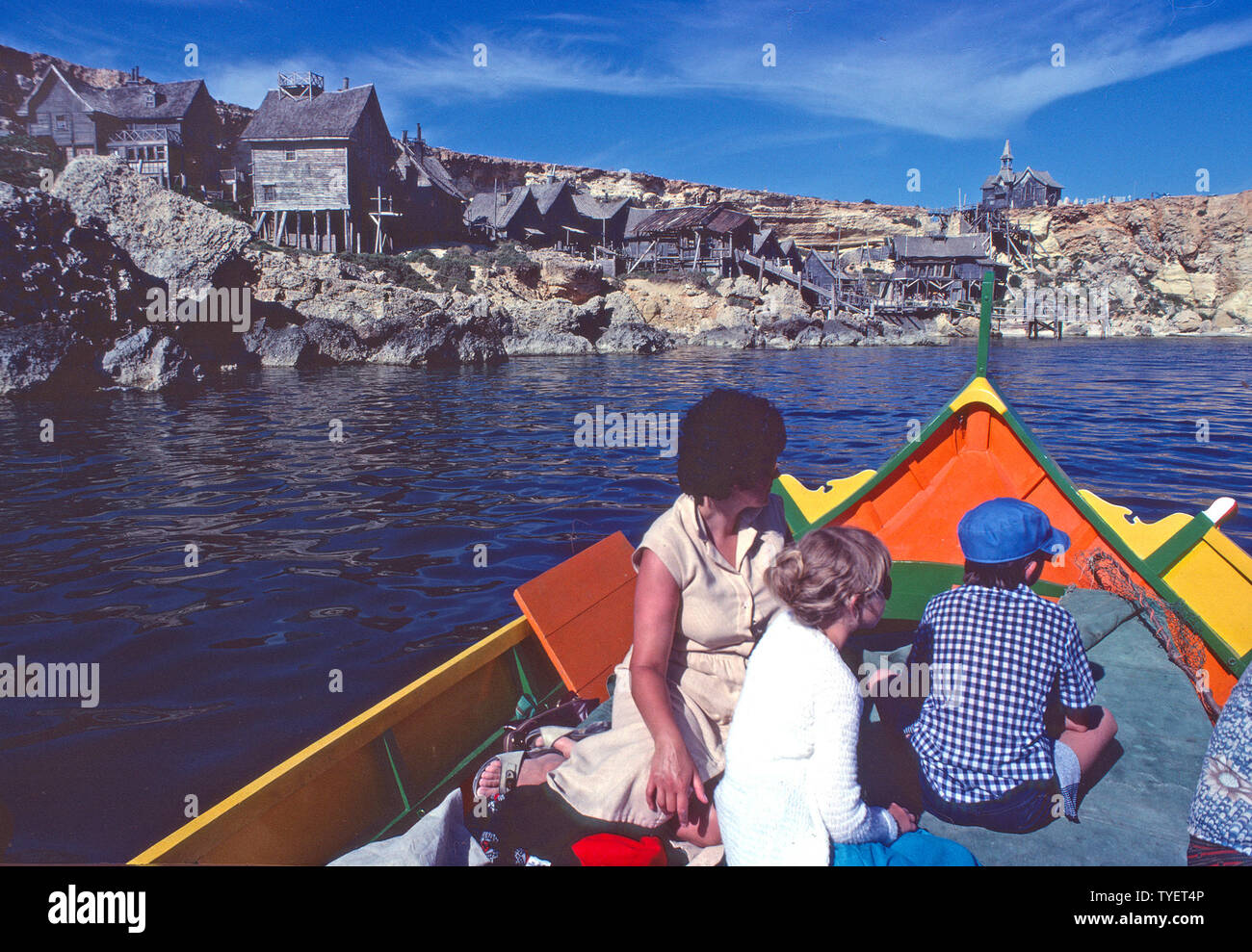 Immagine storica di archivio di madre e bambini britannici anni '80 su Pacchetto vacanze mediterraneo per famiglie a bordo di una colorata barca da pesca maltese All'edificio abbandonato set di film di legno dal Popeye 1980 Filma il modo in cui eravamo ad Anchor Bay Village 80 di Malta Foto Stock