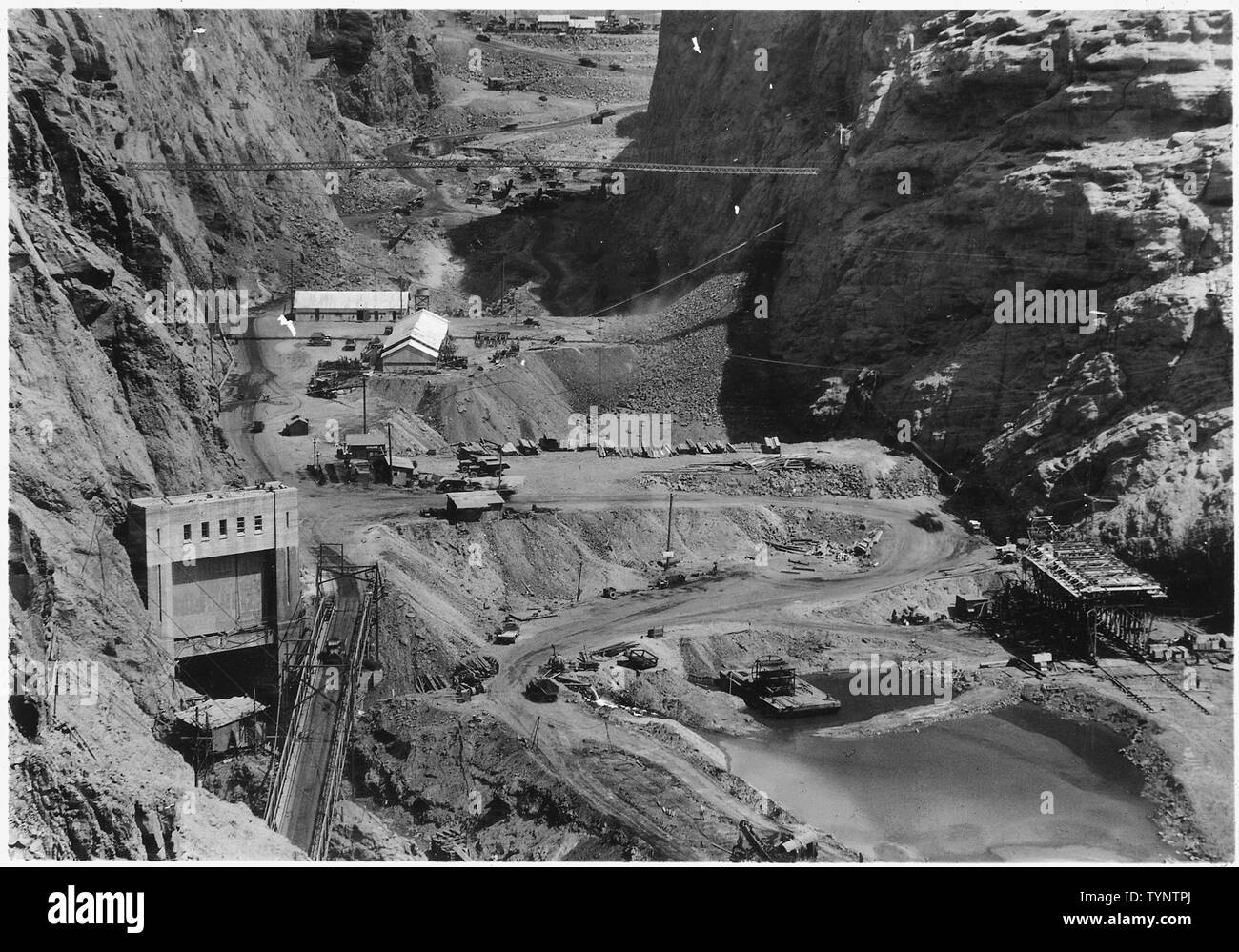 Vista guardando a monte attraverso canyon nero mostra a valle paratoia e rock risacca barriera. Scavo nella sottostruttura dam ed a monte di paratoia visto in background. Costruzione di uffici e negozi visto sulle creste delle paratoie. Stoney struttura di gate al portale di uscita del tunnel di deviazione n. 2 visto in basso a sinistra.; Portata e contenuto: fotografia dal volume due di una serie di album di foto per documentare la costruzione della Diga di Hoover, Boulder City, Nevada. Foto Stock