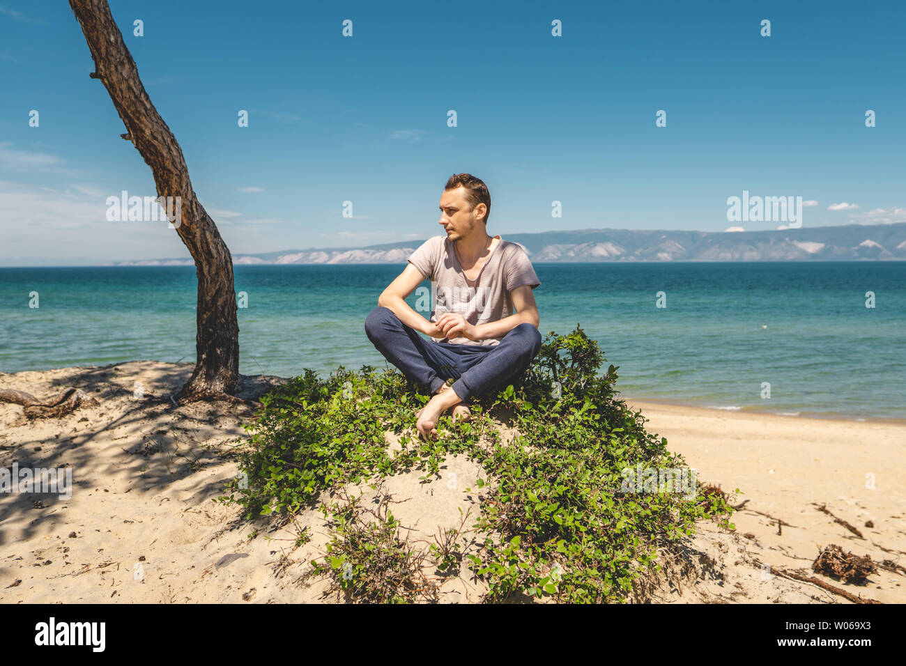 L'uomo viaggiatore rilassante sulla spiaggia di Olkhon island con vista sull'acqua del lago e delle montagne e il cielo blu. La meditazione calma e libertà Foto Stock
