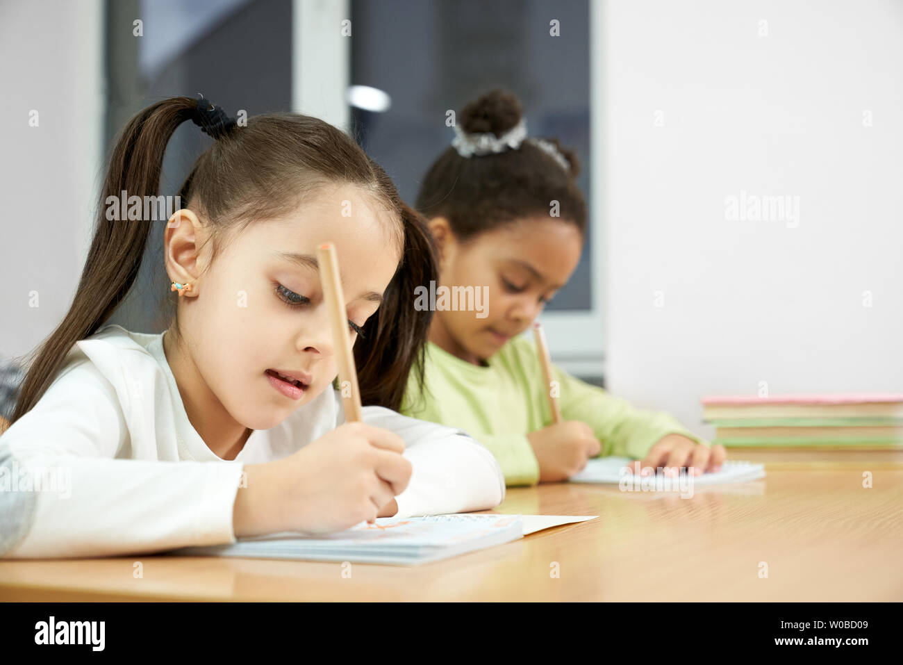 Allegro belle ragazze della scuola primaria in scrittura copybook con matita. Carino, concentrato studentesse seduta in Aula, cercando in notebook, ponendo a lezione. Foto Stock