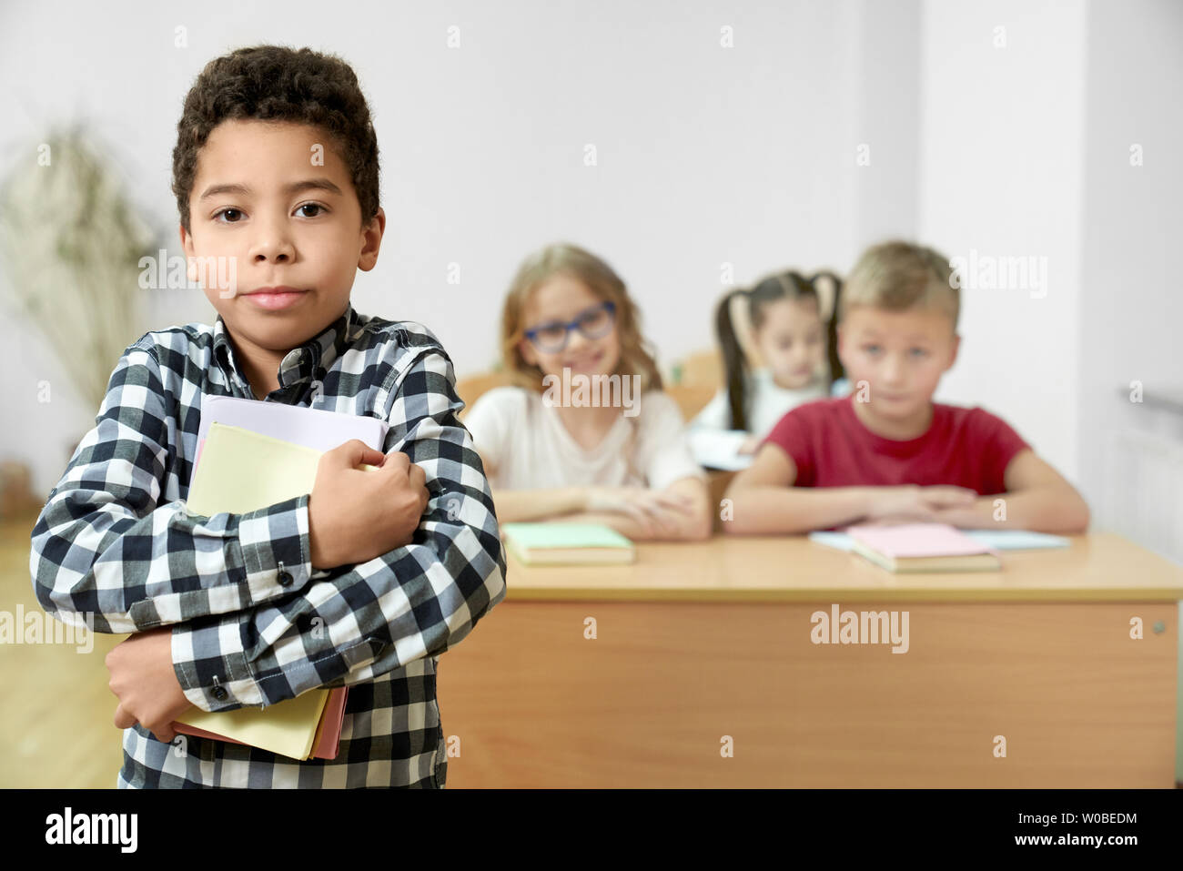 Bello schoolboy in piedi in aula, tenendo in mano libri, guardando la telecamera, in posa. Allegro alunni seduti alla scrivania sullo sfondo. Foto Stock