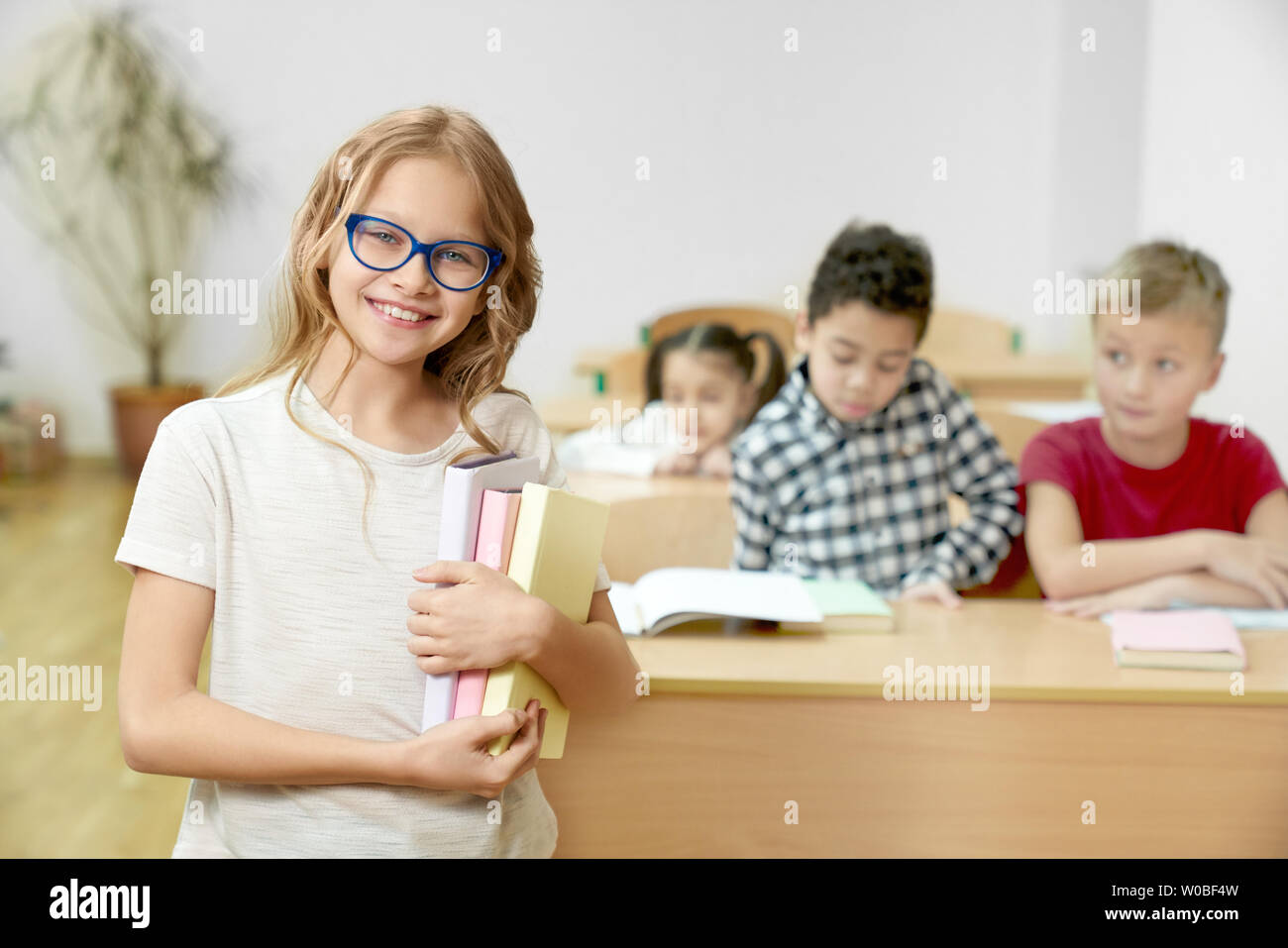 Bello, grazioso, schoolgirl con capelli lunghi in vetri blu libri di contenimento, in piedi in aula. Felice, positivo adolescente che studiano nella scuola primaria guardando la telecamera, in posa. Compagni di classe su sfondo. Foto Stock