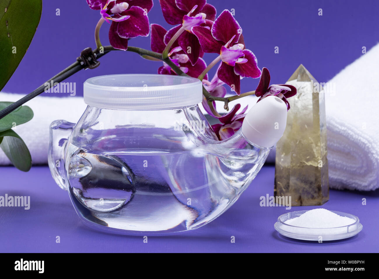 Neti Pot, soluzione salina, fiori di orchidea, arrotolato asciugamani bianchi e lucidato Quarzo Citrino punto su sfondo viola. Lavaggio del seno. Irrigazione nasale. Foto Stock