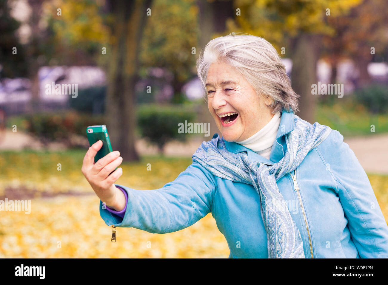 Vista di un smilling ed espressive piuttosto alti donna guardando al suo telefono su foglie di giallo nella parte anteriore di un alberi con colori autunnali. Foto Stock