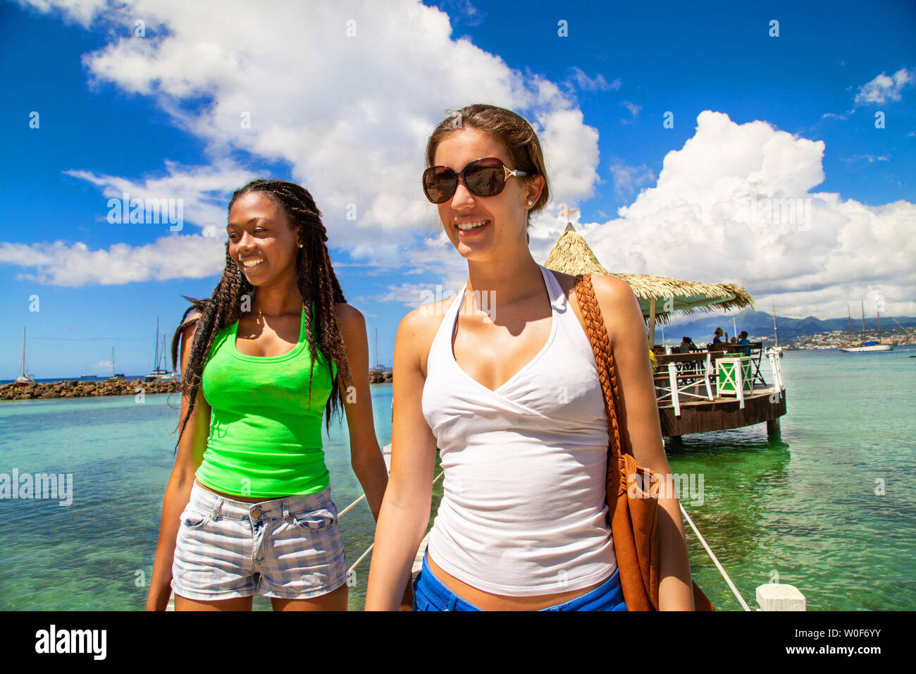 2 amiche sorridenti a camminare su una passerella proveniente da un bar su palafitte su una spiaggia caraibica in un paesaggio paradisiaco. Foto Stock