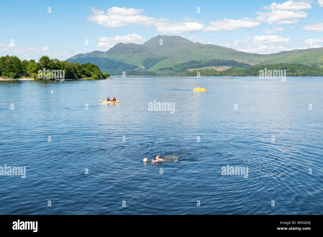 Luss, Loch Lomond Scozia, Regno Unito - 27 Giugno 2019: Regno Unito meteo - una cartolina impostazione come la gente a prendere alle acque di raffreddamento del Loch Lomond su di una teglia calda giornata Luss, al di sotto del picco di distintivo di Ben Lomond Credito: Kay Roxby/Alamy Live News Foto Stock