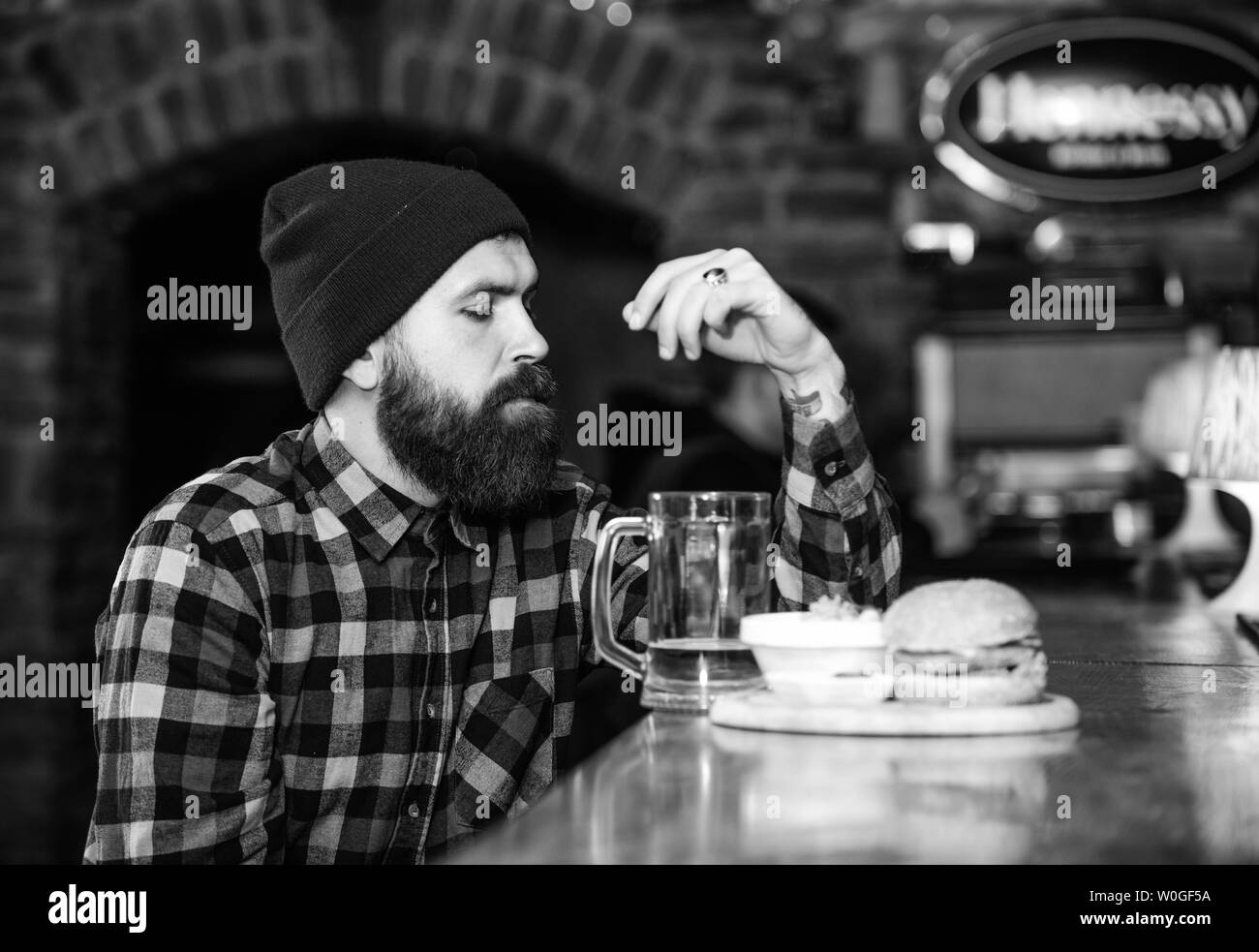 Ad alto contenuto calorico snack. Hipster rilassante al pub. Pub è un luogo rilassante per avere bere e rilassarsi. Uomo con barba bere birra mangiare hamburger menu. Pranzare in un pub. Brutale parigamba uomo barbuto sedersi al banco bar. Foto Stock