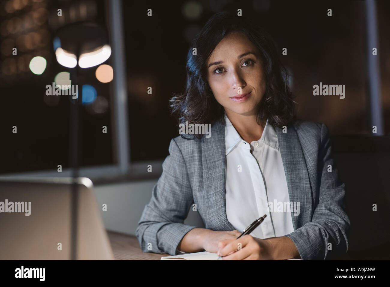 Ritratto di una giovane imprenditrice seduto alla sua scrivania scrivere note e utilizzando un computer portatile mentre lavoro straordinario in un ufficio a tarda sera Foto Stock