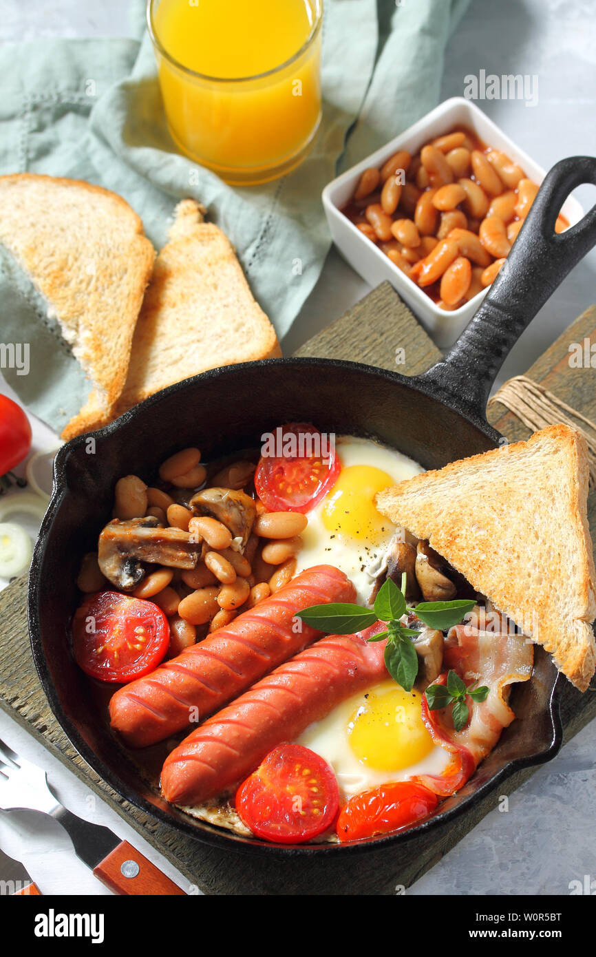 Prima colazione in inglese le uova strapazzate con pancetta, fagioli, funghi, salsicce, pomodori e succo di luce sul tavolo di pietra. Foto Stock