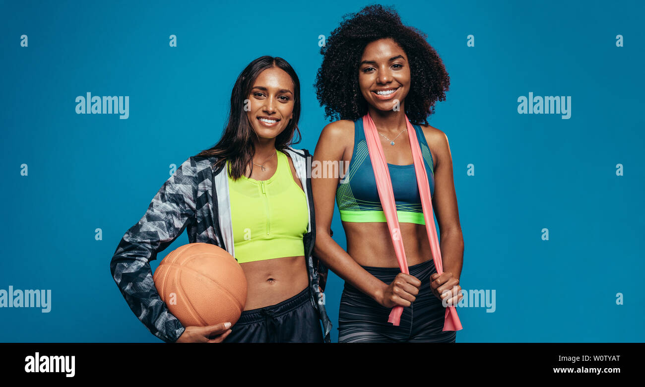 Due giovani donne in piedi insieme con la pallacanestro e resistenza band su sfondo blu. Razza mista amici di sesso femminile in abbigliamento sportivo guardando ca Foto Stock