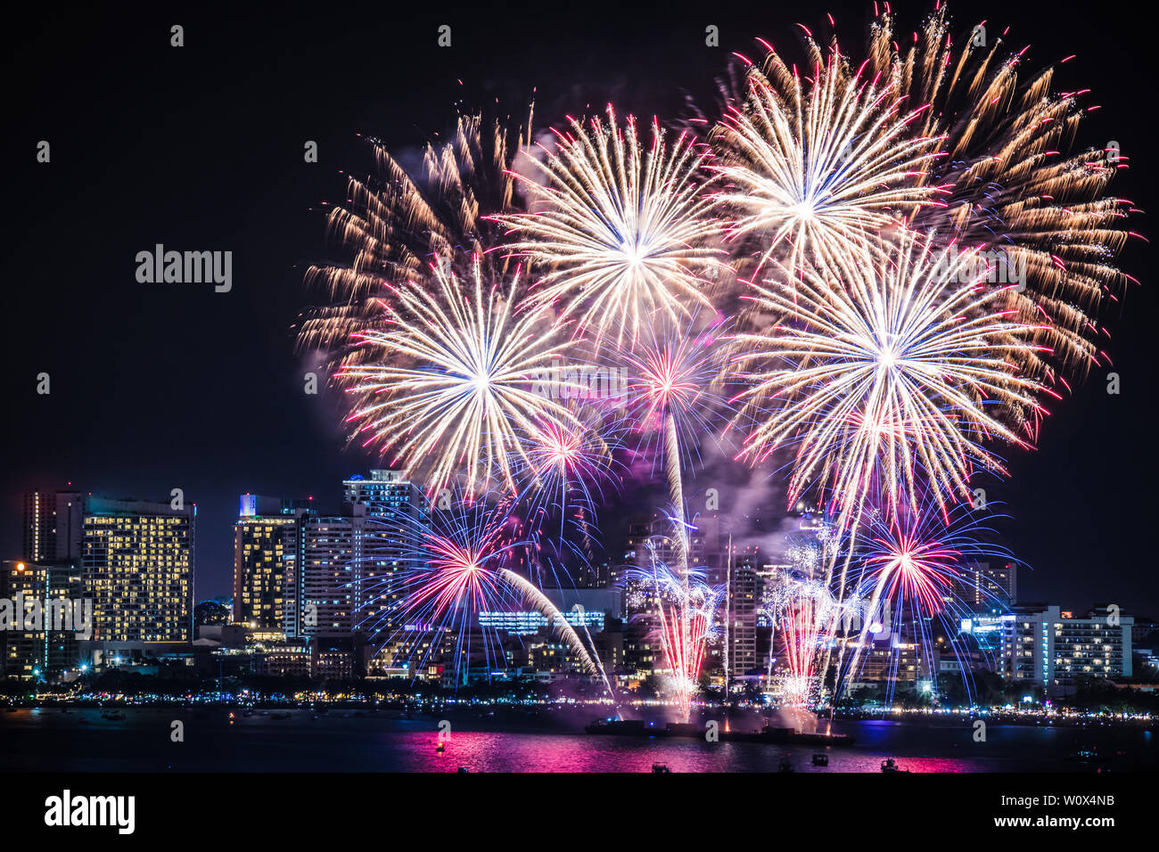 Vero e proprio festival dei fuochi d'artificio in cielo per la celebrazione di notte con vista della città a sfondo e barca galleggiante sul mare al primo piano a lato della costa Foto Stock