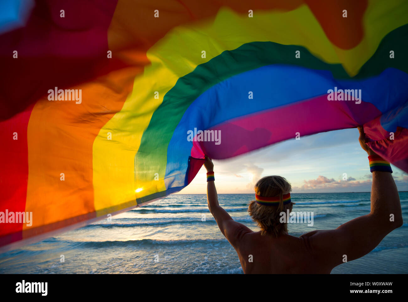 Silhouette di uomo con capelli biondi indossa LGBTQI archetto orgoglio tenendo un svolazzanti bandiera arcobaleno sulla spiaggia di sunrise Foto Stock