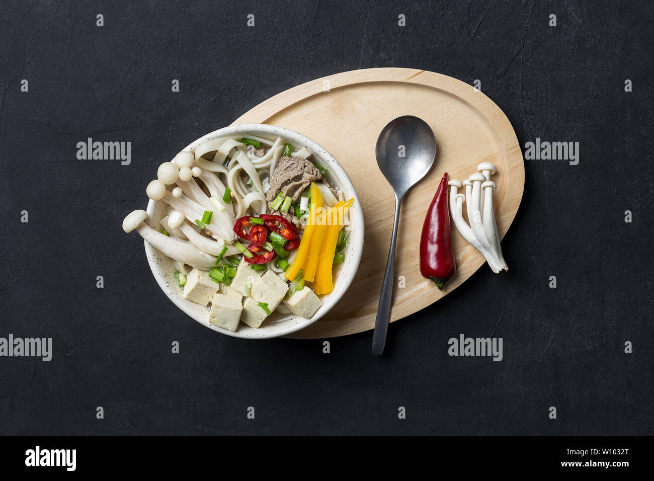 Giapponese calda zuppa di noodle con carne di manzo, giallo paprika, funghi, cipolla verde, tofu e peperoncino su sfondo nero con il cucchiaio di legno e vassoio. Concetto di Foto Stock