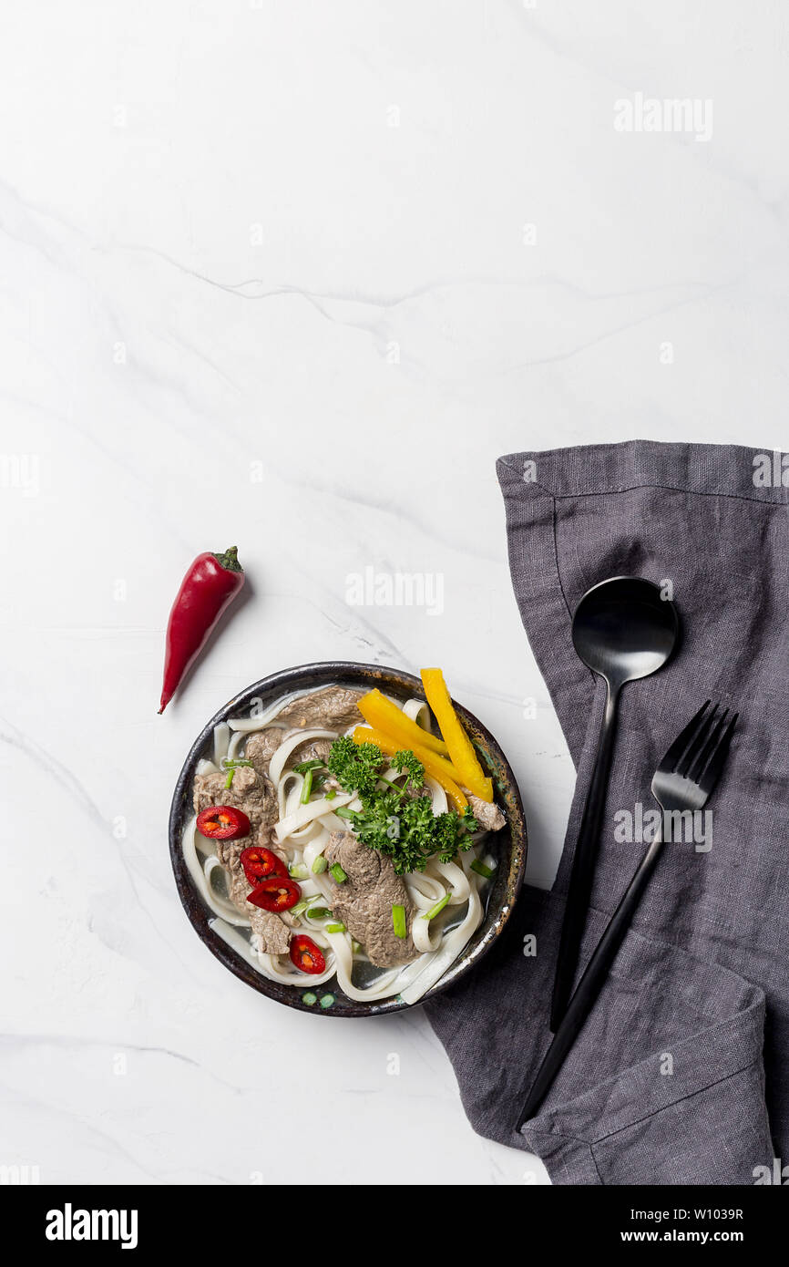 Asian zuppa di noodle, la carne e le verdure nella ciotola con il tovagliolo grigio e posate su sfondo bianco. Verticale in alto vista, lo spazio libero per il testo Foto Stock