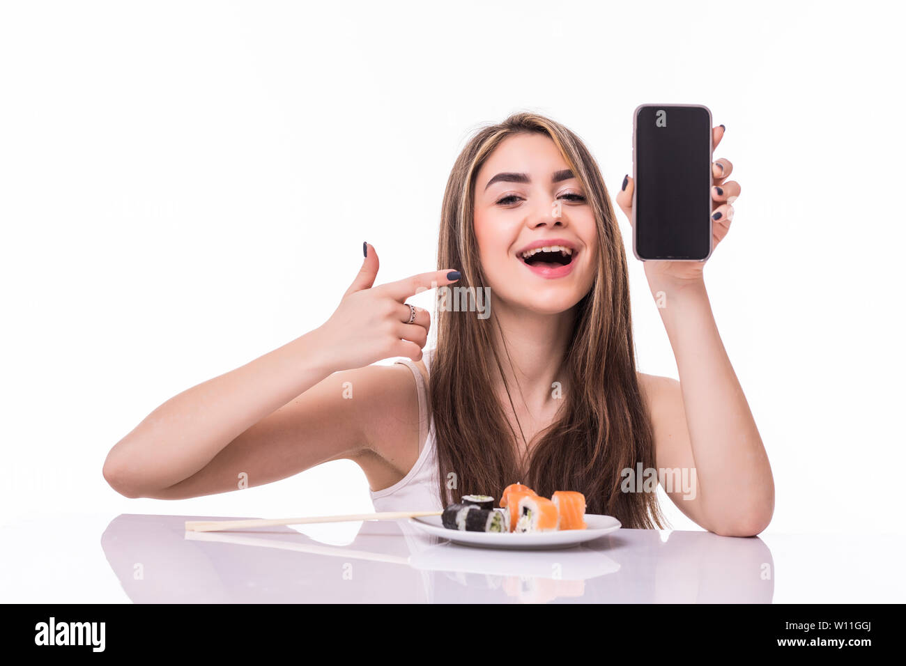 Sorridente giovane donna asiatica di mangiare sushi al tavolo isolate su sfondo bianco, mostra schermo vuoto del telefono mobile Foto Stock