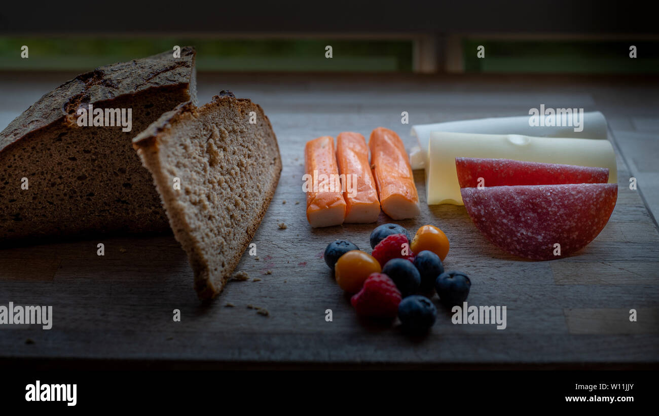 Un low-carb , proteina-ricco pasto costituito da formaggio svizzero, salame; bacche, fake pesce e pane di segale sul piatto di legno da una finestra Foto Stock