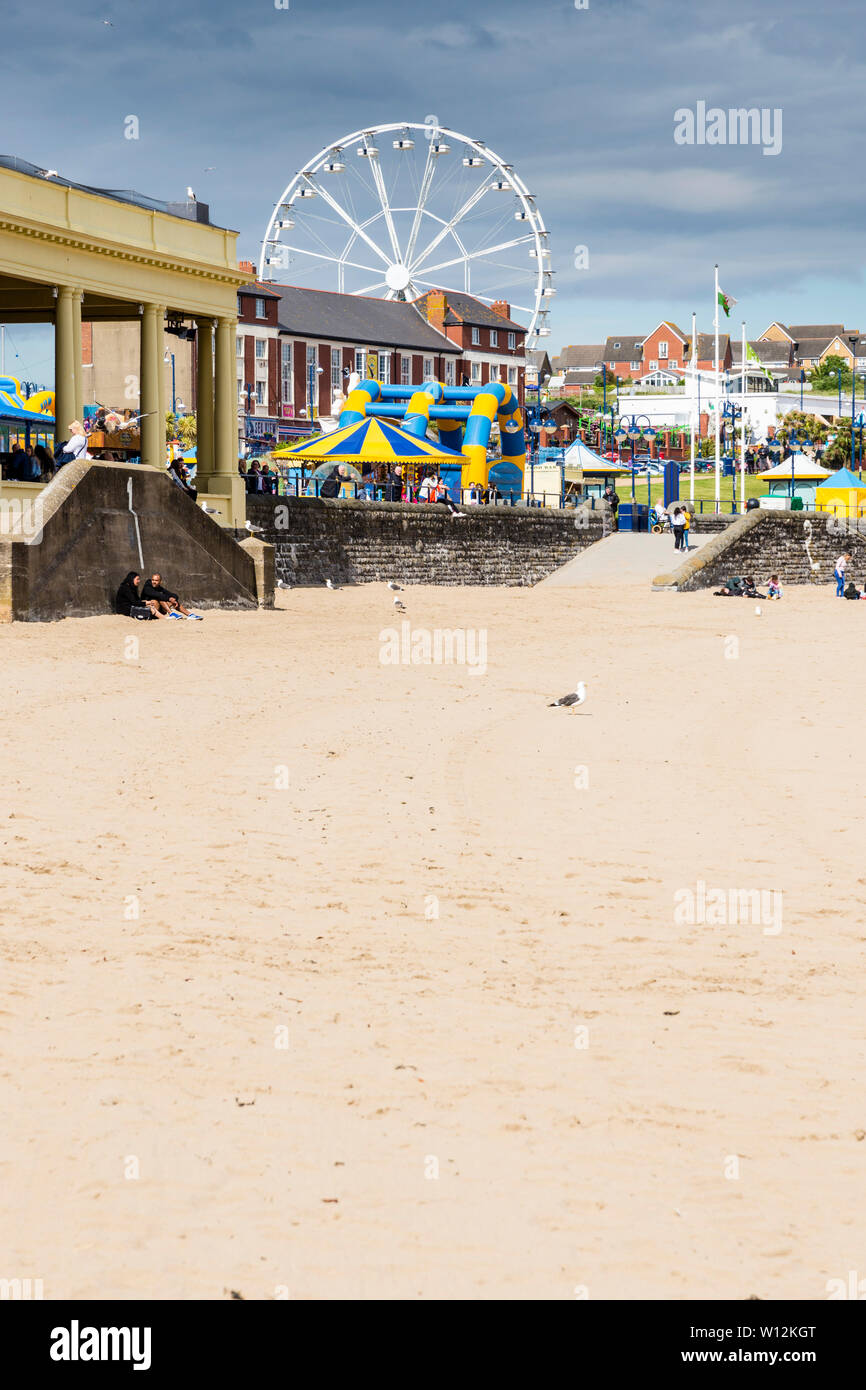 La spiaggia di sabbia fine e ruota panoramica Ferris a Barry Island sono illuminate dal sole e si stagliano contro le nuvole scure nel cielo con un inizio di giornata estiva Foto Stock