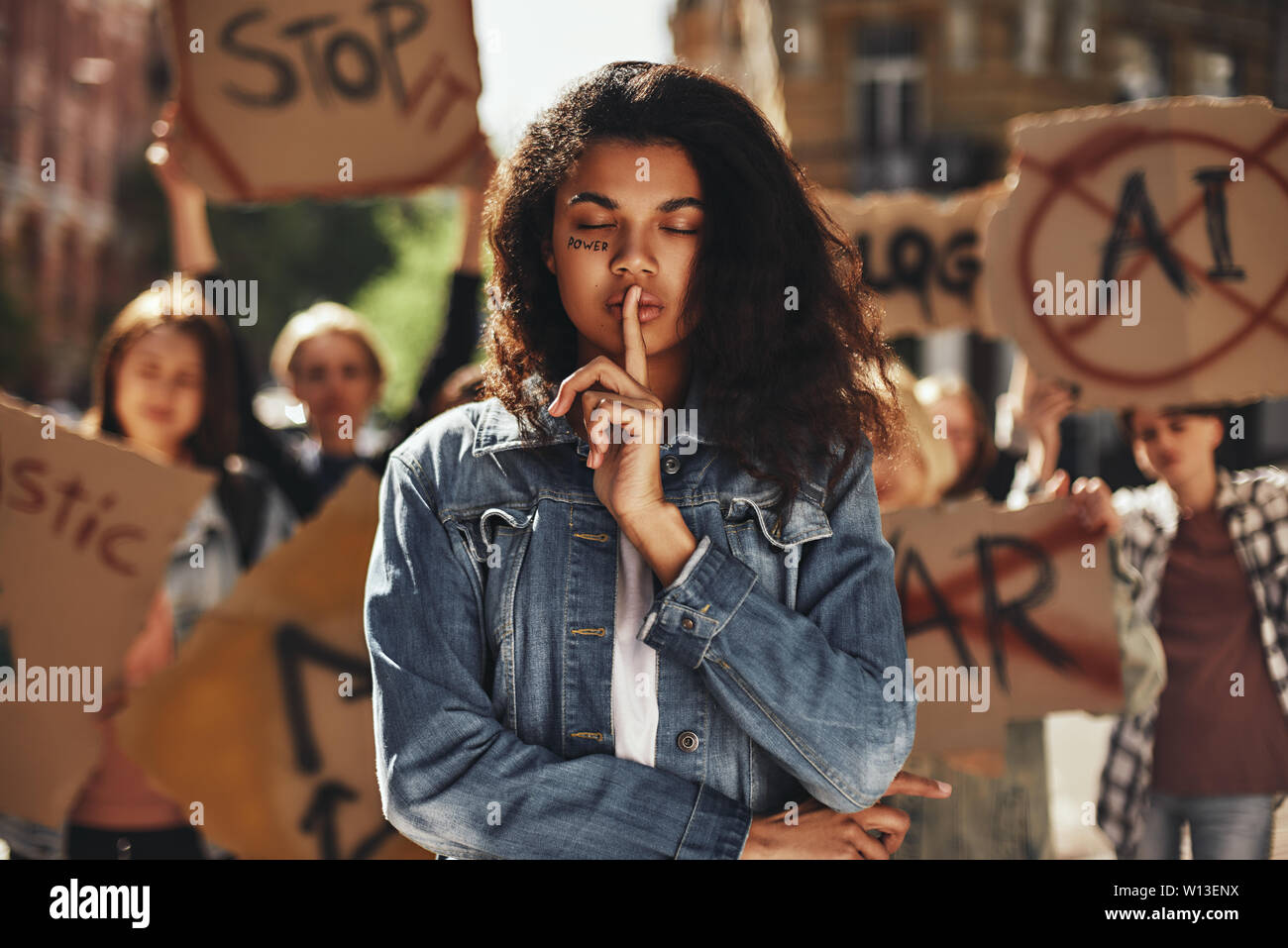 Giovani afro american donna con potenza di parola scritta sul suo viso e con gli occhi chiusi che mostra segni di silenzio durante una protesta con un gruppo di attivisti femmina all'aperto sulla strada. Diritti dell'uomo. La dimostrazione. Forza delle donne Foto Stock