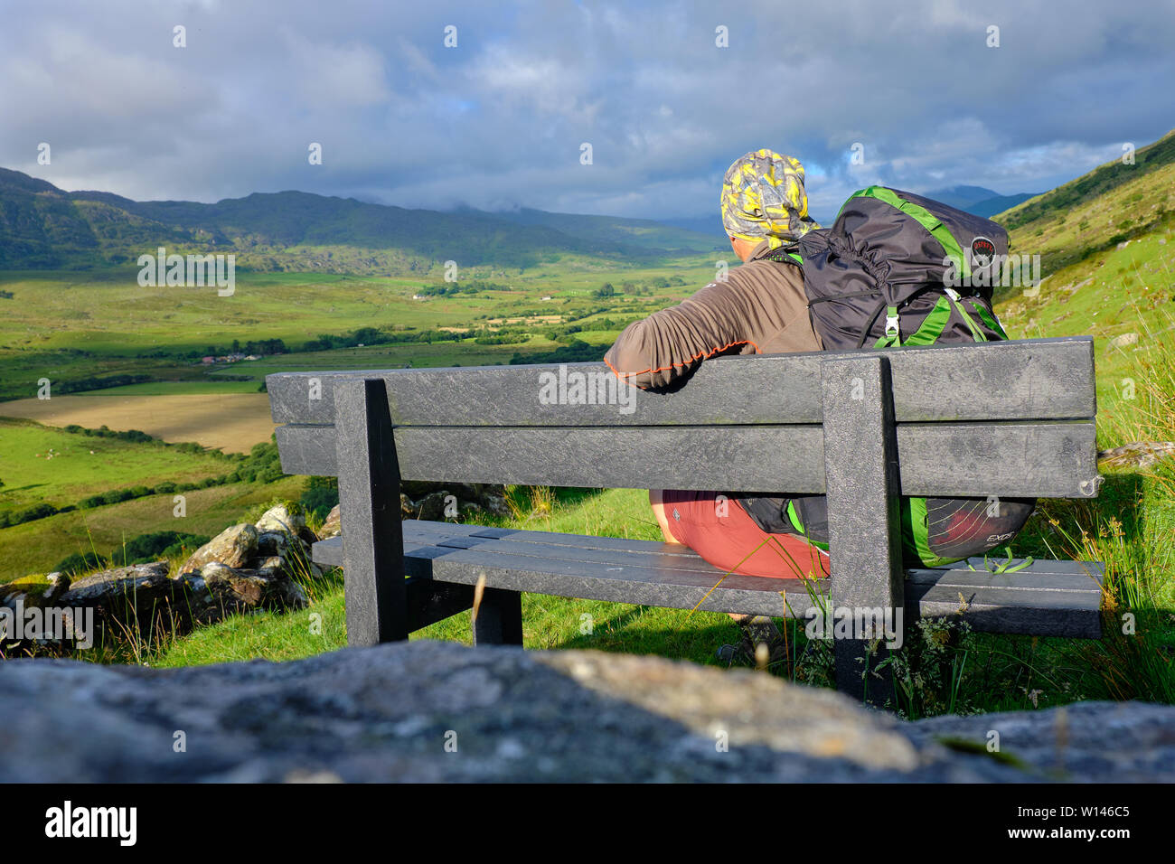 Escursionista maschio indossa uno zaino sul modo di Kerry seduta su una panchina a riposare guardando al pascolo verde vista. Foto Stock